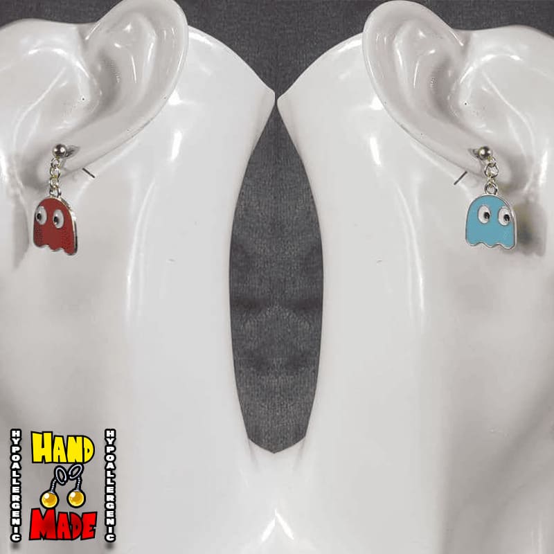 A pair of handcrafted metal earrings, one resembling a blue Pac-Man ghost and the other a red Pac-Man ghost, displayed on a white earring stand.