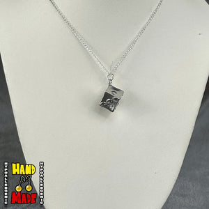 A silver-colored cube-shaped metal charm on a silver chain necklace, displayed on a mannequin.