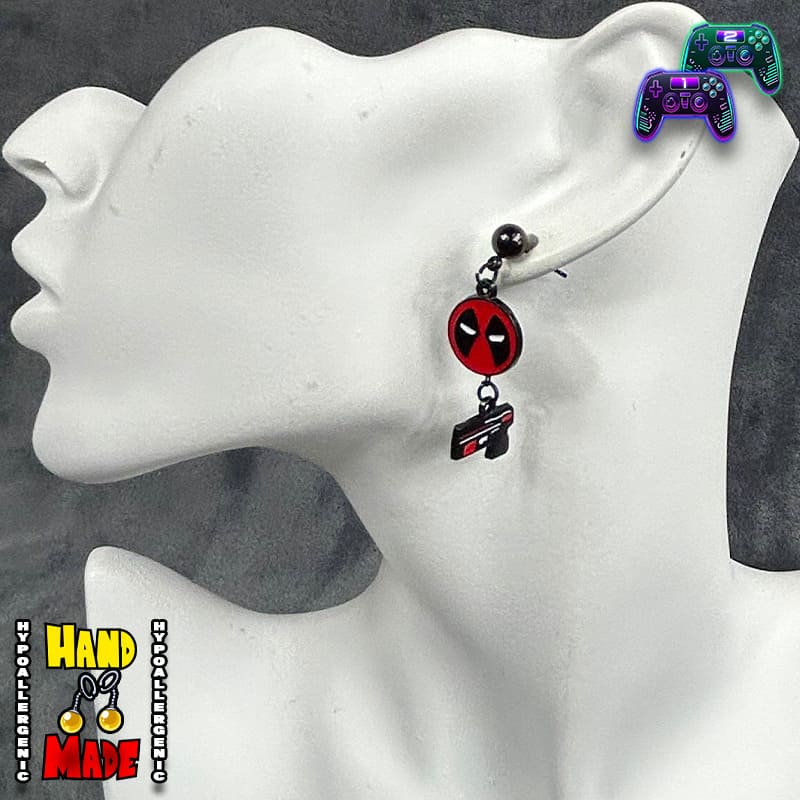 A pair of Deadpool-themed earrings displayed on a mannequin head. One earring features a red and black gun charm, and the other has a solid black gun charm, both designed to dangle behind a mask.