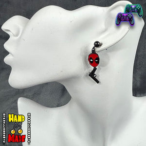 A pair of Deadpool-themed earrings displayed on a mannequin head. One earring features a red and black gun charm, and the other has a solid black gun charm, both designed to dangle behind a mask.