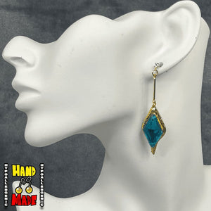 A pair of turquoise diamond cut earrings, hanging from a gem dropped post, with gold accents, displayed on a mannequin ear.