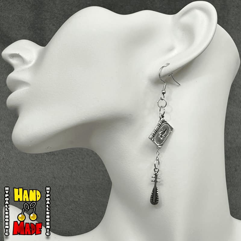 A pair of silver-tone earrings featuring a book charm and a hanging cudgel charm, displayed on a mannequin ear.