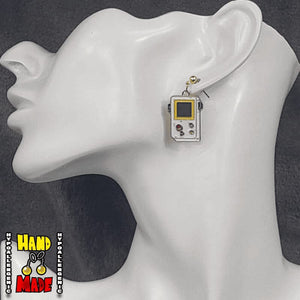 A pair of gaming earrings designed to look like a retro Gameboy with joystick, hanging from a gold dropped post, displayed on a mannequin ear.