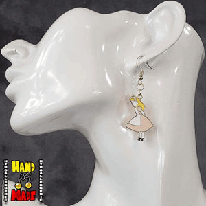 A pair of handmade gaming earrings, featuring an oversized enamel charm with a character in a pink dress, resembling Alice from 'Alice in Wonderland', displayed on a mannequin ear.