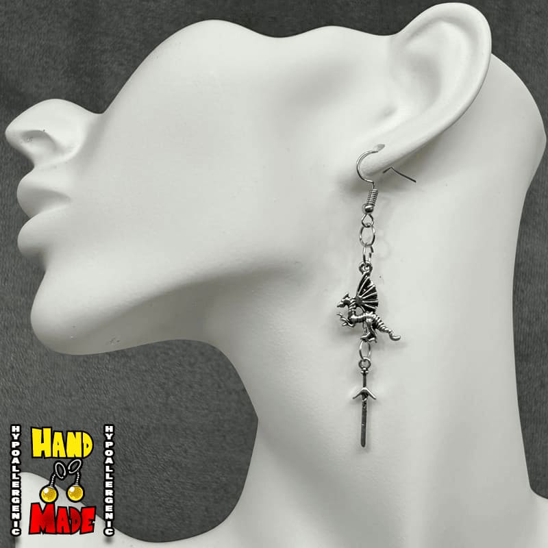 A pair of silver metal dragon charms, with a hanging sword charm, displayed on a mannequin ear.