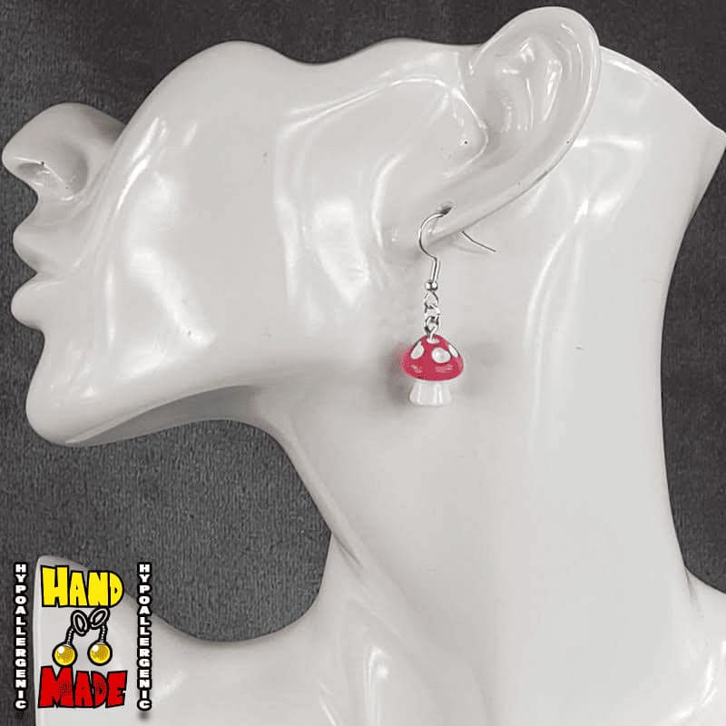 A pair of bright pink and white plastic mushroom charm earrings, hanging from a silver French hook, displayed on a white mannequin head.