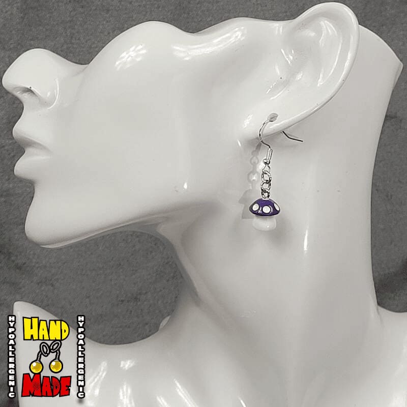 A pair of purple and white plastic mushroom charm earrings, hanging from a silver French hook, displayed on a white mannequin head.