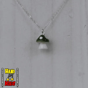 A dark green and white plastic mushroom charm, hanging from a silver chain, displayed on a white board.