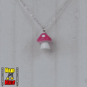 A hot pink and white plastic mushroom charm, hanging from a silver chain, displayed on a white board.