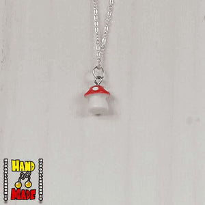 A red and white plastic mushroom charm, hanging from a silver chain, displayed on a white board.