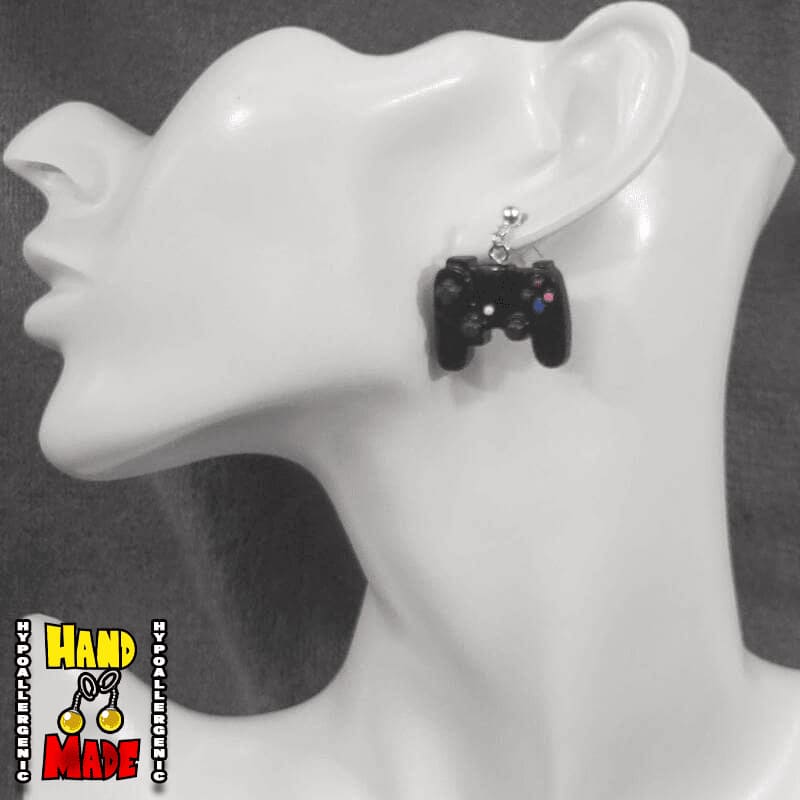 A pair of black plastic earrings designed to resemble X-Box controllers, hanging from a silver dropped post, displayed on a mannequin head.
