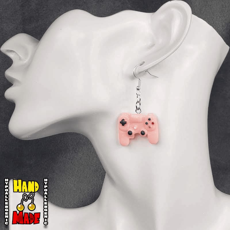 A pair of pink plastic earrings designed to resemble X-Box controllers, hanging from a silver French hook, displayed on a mannequin head.