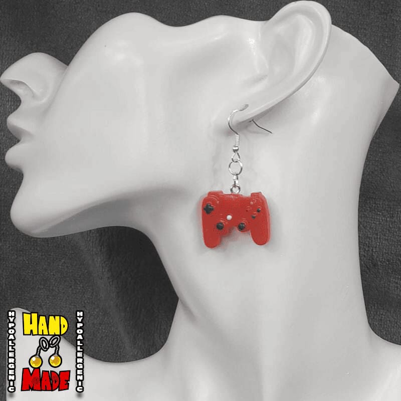 A pair of red plastic earrings designed to resemble X-Box controllers, hanging from a silver French hook, displayed on a mannequin head.