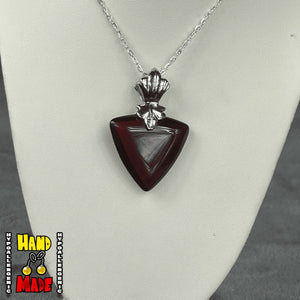 A red triangle-heart-shaped pendant, with a silver chain and ornate detailing, displayed on a white mannequin stand.