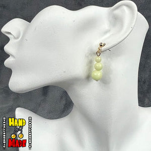 A pair of yellow beaded earrings, featuring a triple-ball stack design, dangling from a gold dropped post, displayed on a mannequin ear.