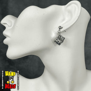 A pair of silver-colored cube earrings on a mannequin head, with the earrings designed to resemble items from the anime series Jujutsu Kaisen.